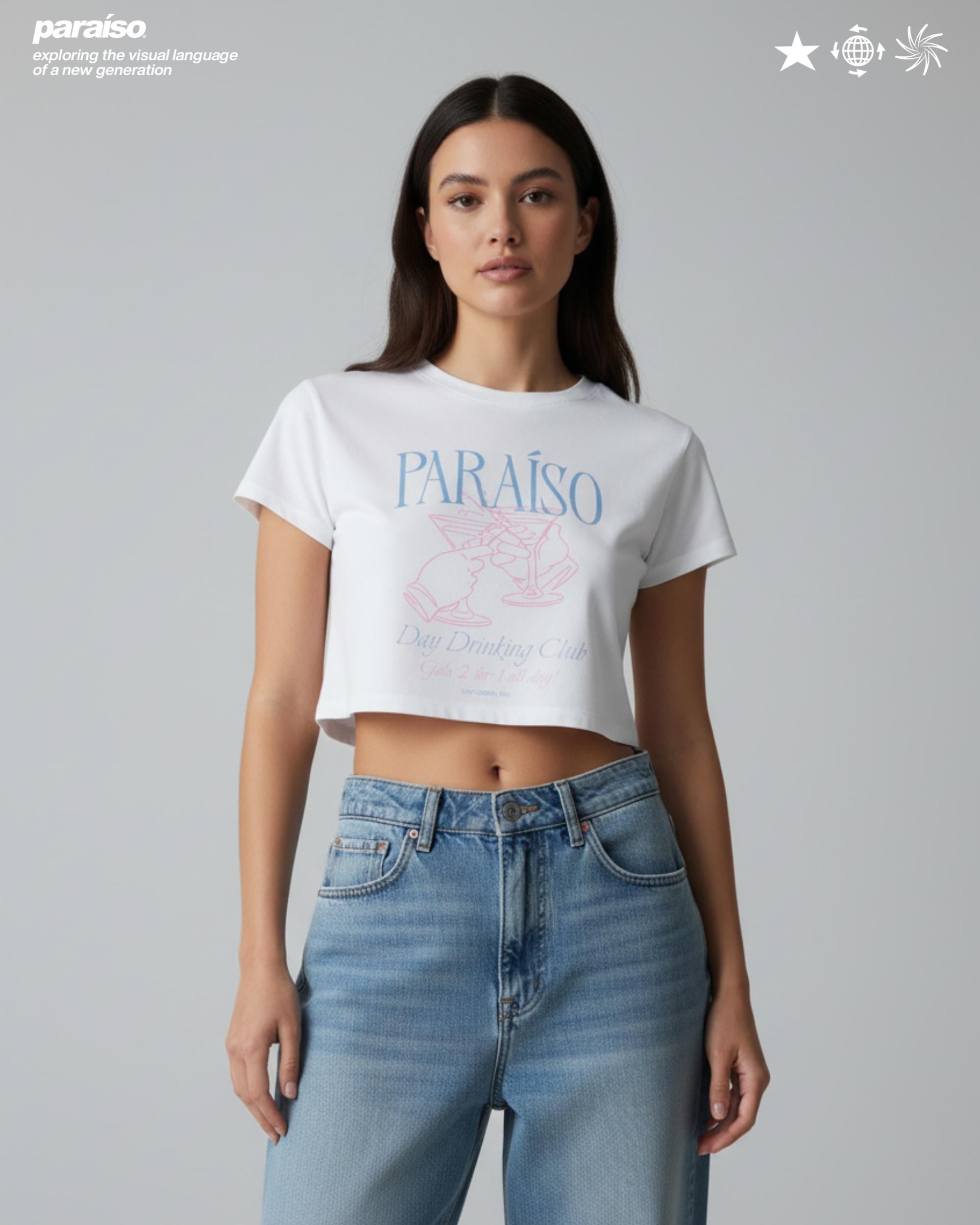 White crop top with the word 'PARAISO' in light blue, and a pink graphic of two figures toasting cocktails. Below the graphic is the text 'Day Drinking Club'. The model is wearing light blue denim jeans.